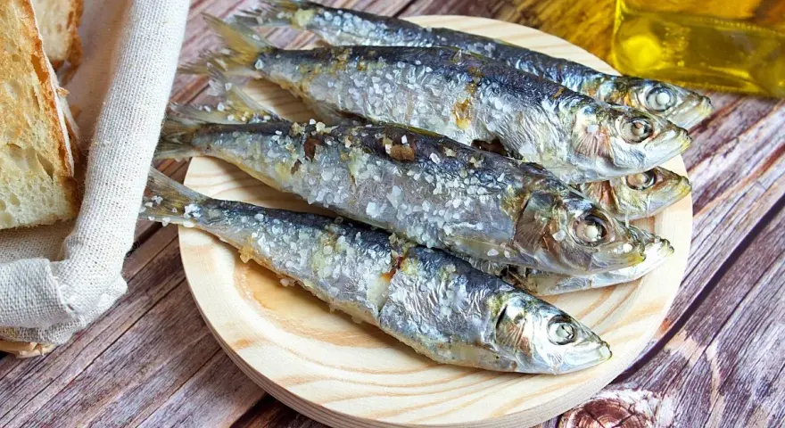 Cinco sardinas al horno, saladas y doradas, reposan en un plato de madera junto a pan rústico y una botella de aceite.