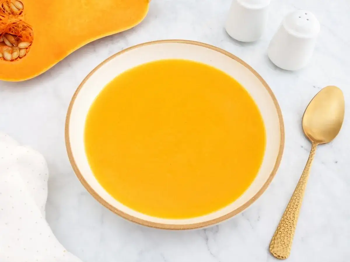 Un tazón de cremoso pure de calabaza junto a una calabaza cortada, una cuchara dorada y saleros.