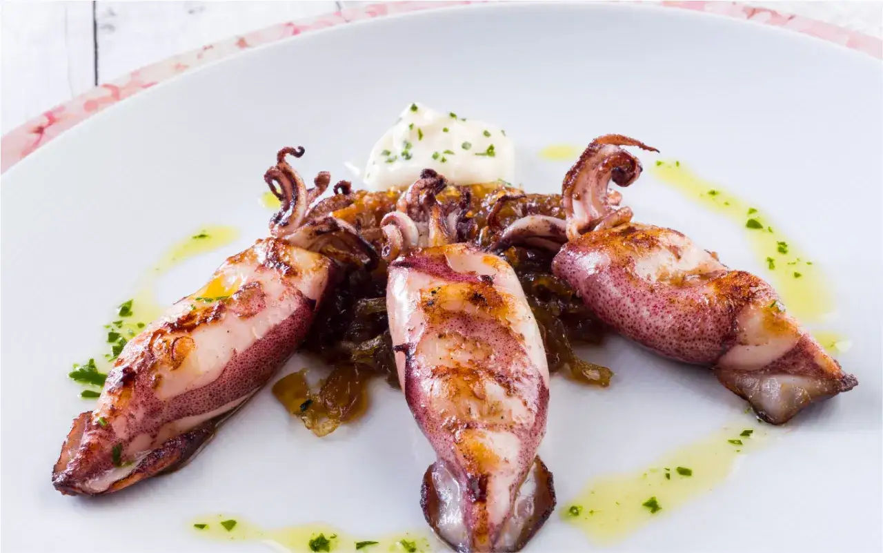 Tres calamares a la plancha, dorados y jugosos, servidos sobre una cama de cebolla caramelizada y acompañados de alioli y aceite de oliva con perejil.
