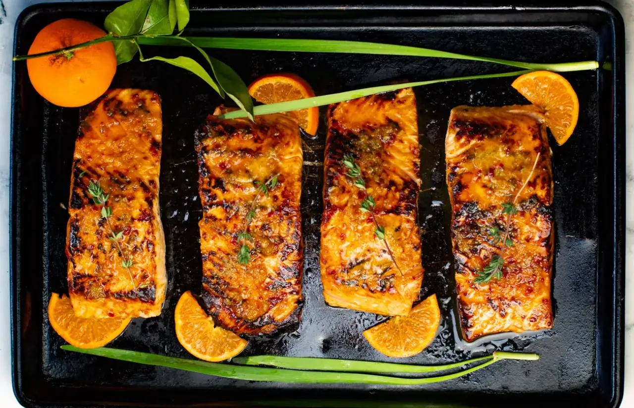 Cuatro filetes de salmón a la naranja glaseados y dorados, adornados con ramitas de tomillo, gajos de naranja y cebolleta verde.