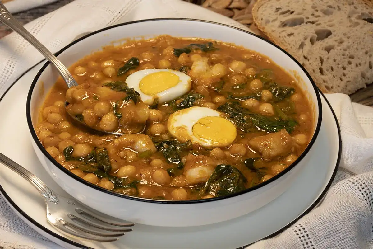 Un plato humeante de garbanzos con espinacas y trozos de huevo duro, listo para disfrutar con pan rústico.