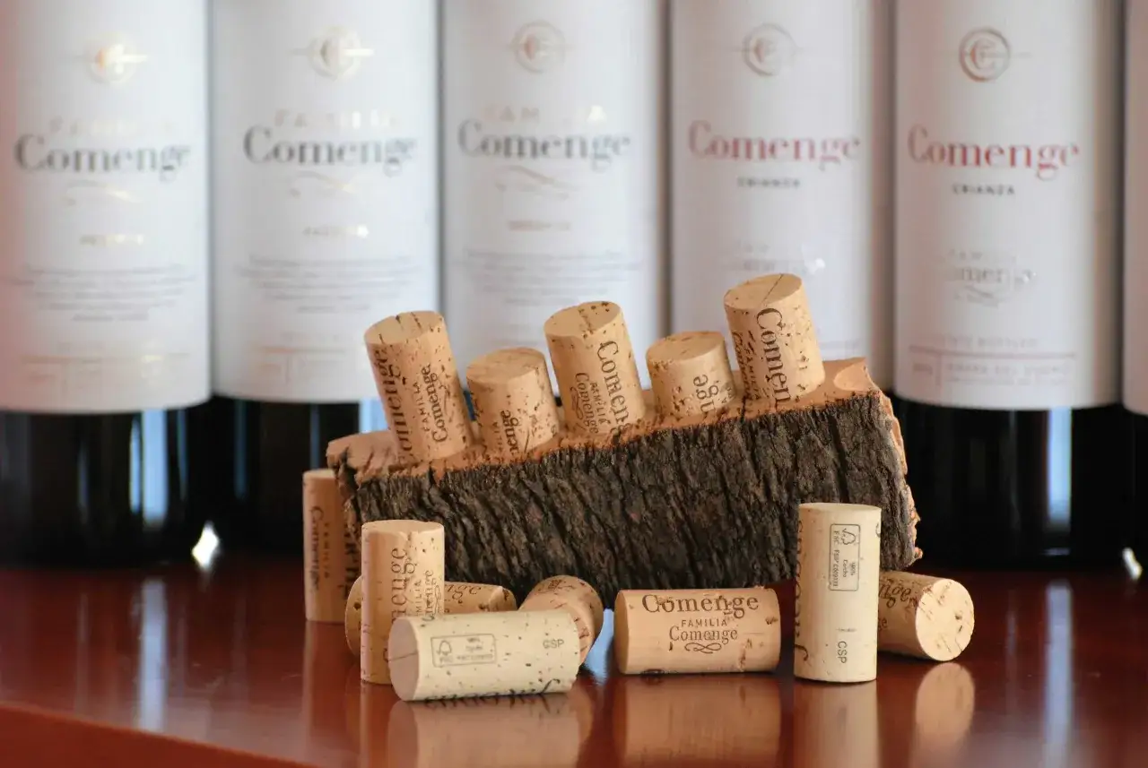 Tapones de corcho, que nos recuerdan de donde viene el corcho, apoyados sobre un trozo de corteza, con botellas de vino al fondo.