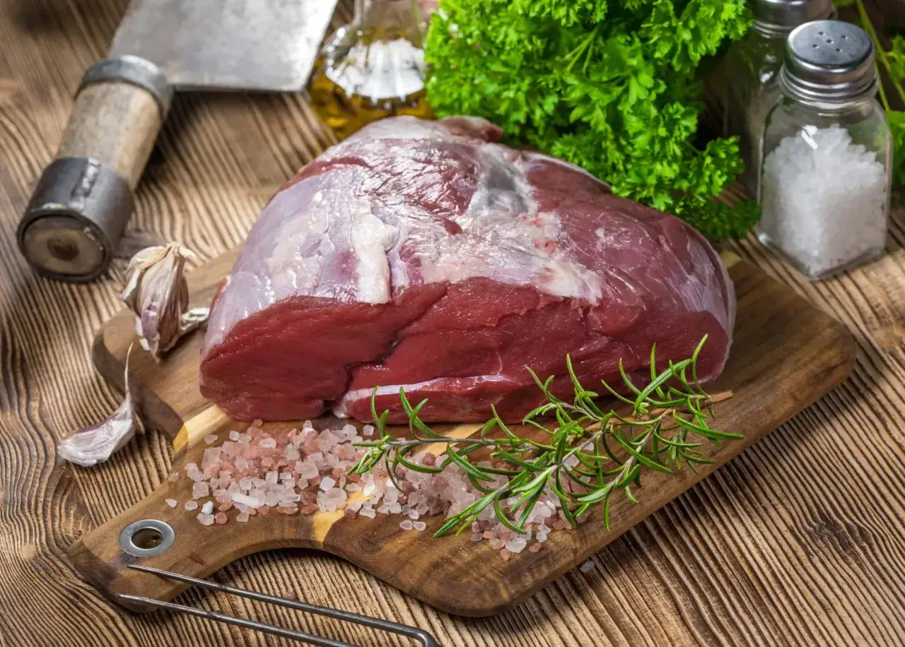 Un trozo de carne roja, que es la ternera, reposa sobre una tabla de madera con sal rosada, romero, ajo y perejil.