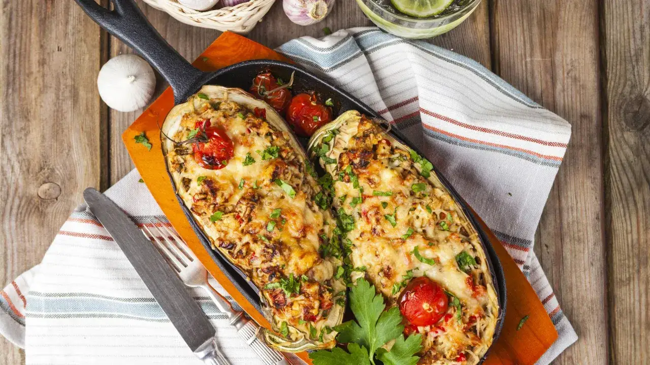 Dos mitades de berenjenas rellenas, gratinadas con queso y adornadas con tomates cherry y perejil, servidas en una sartén de hierro fundido.