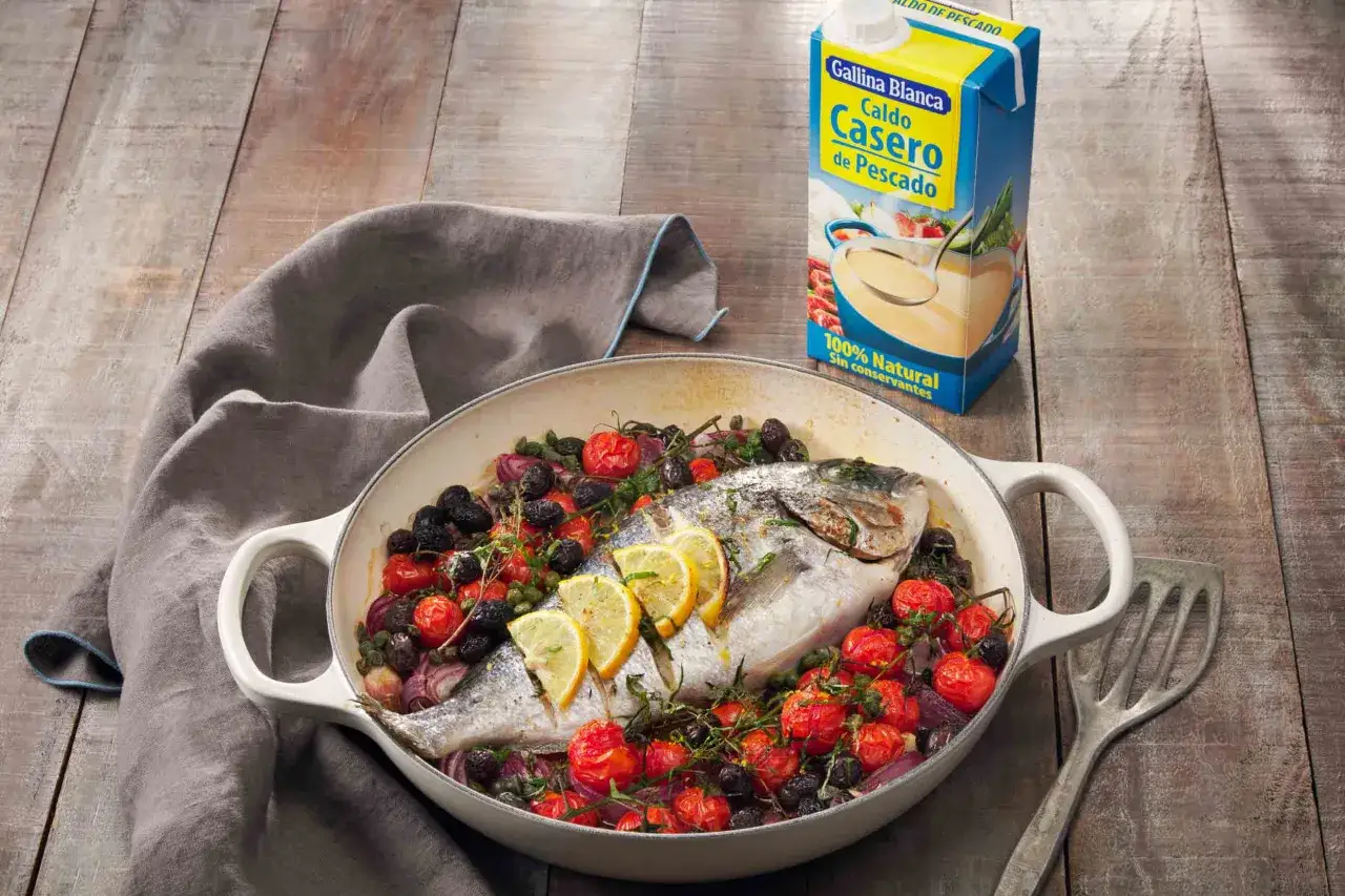 Dorada al horno con patatas, tomates cherry, aceitunas negras y rodajas de limón en una sartén blanca sobre una mesa de madera.
