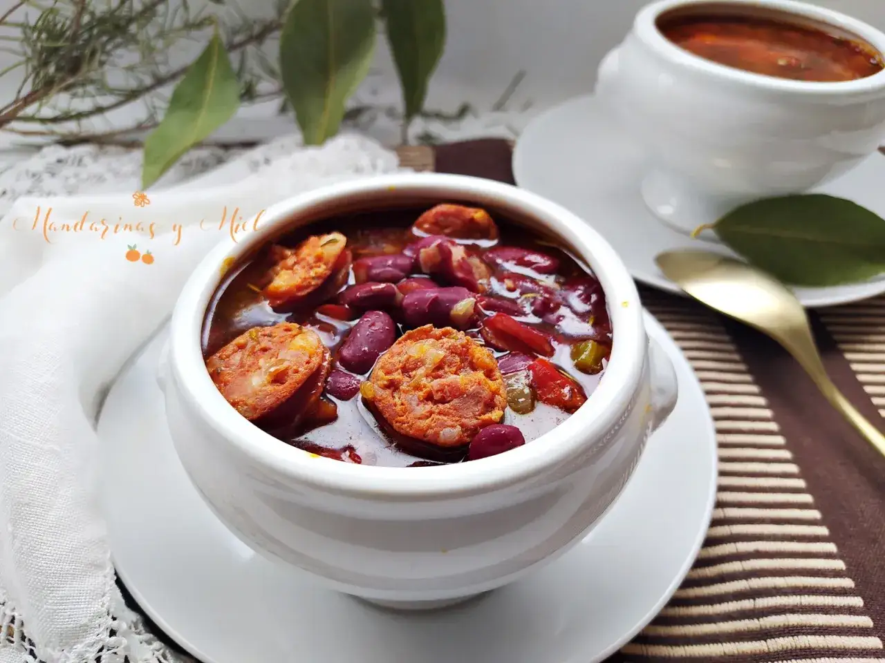 Un reconfortante plato de alubias con chorizo, servido en un cuenco blanco, con hojas verdes y una cuchara dorada.