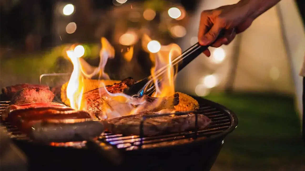 Carne y maíz asándose en una barbacoa, con llamas vibrantes. Una mano usa pinzas para voltear la comida, capturando la esencia de qué es barbacoa.