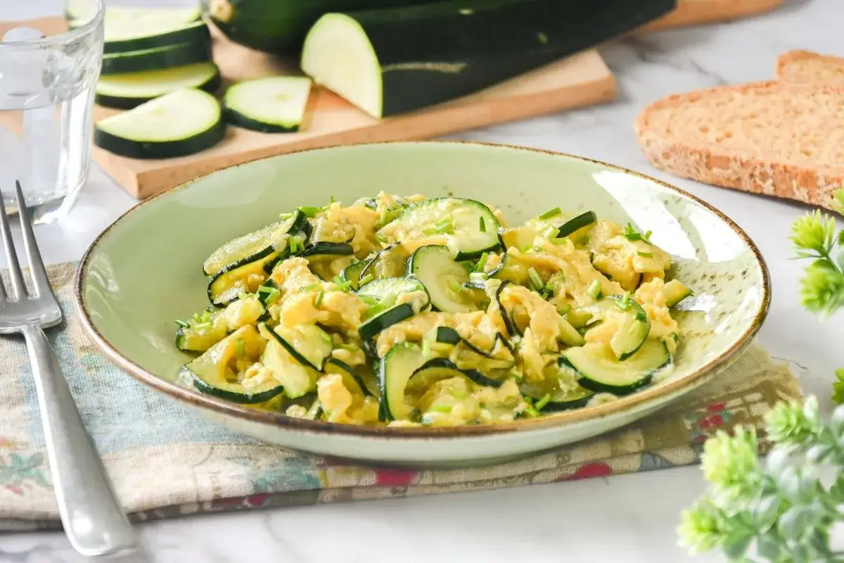 Plato de calabacín revuelto con huevo y cebollino, ideal para recetas con calabacín. Acompañado de pan tostado y un vaso de agua.