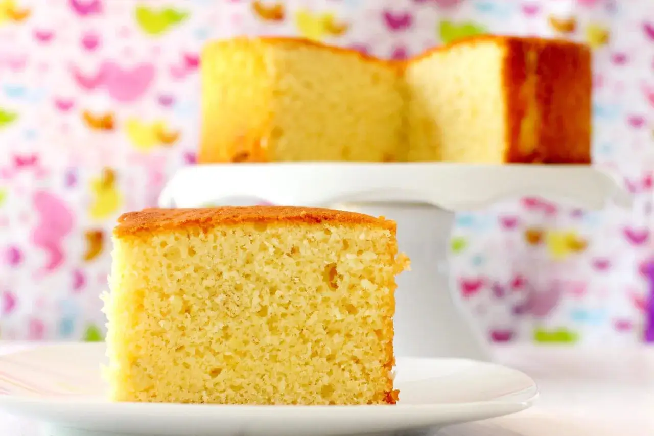 Un trozo de bizcocho de yogur esponjoso en un plato blanco, con otro bizcocho en un soporte de tarta al fondo.