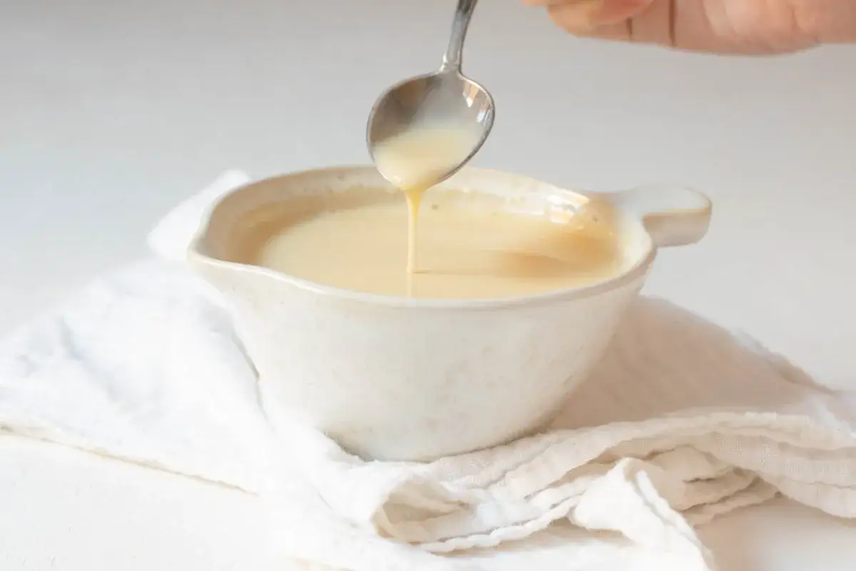 Cuchara vertiendo crema inglesa en un cuenco blanco sobre tela arrugada.