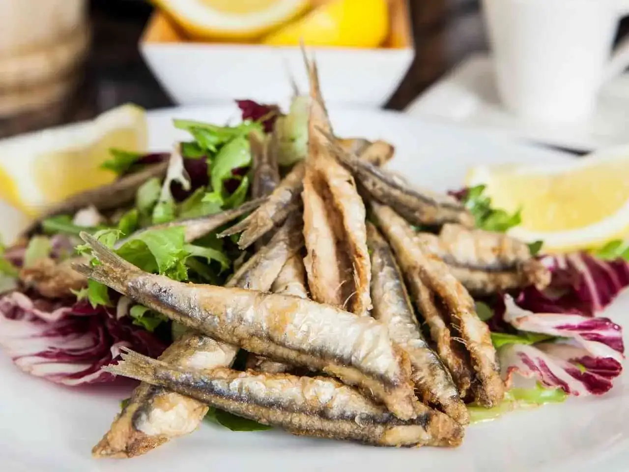 Montón de boquerones fritos crujientes sobre una cama de lechugas variadas y radicchio, acompañados de gajos de limón.