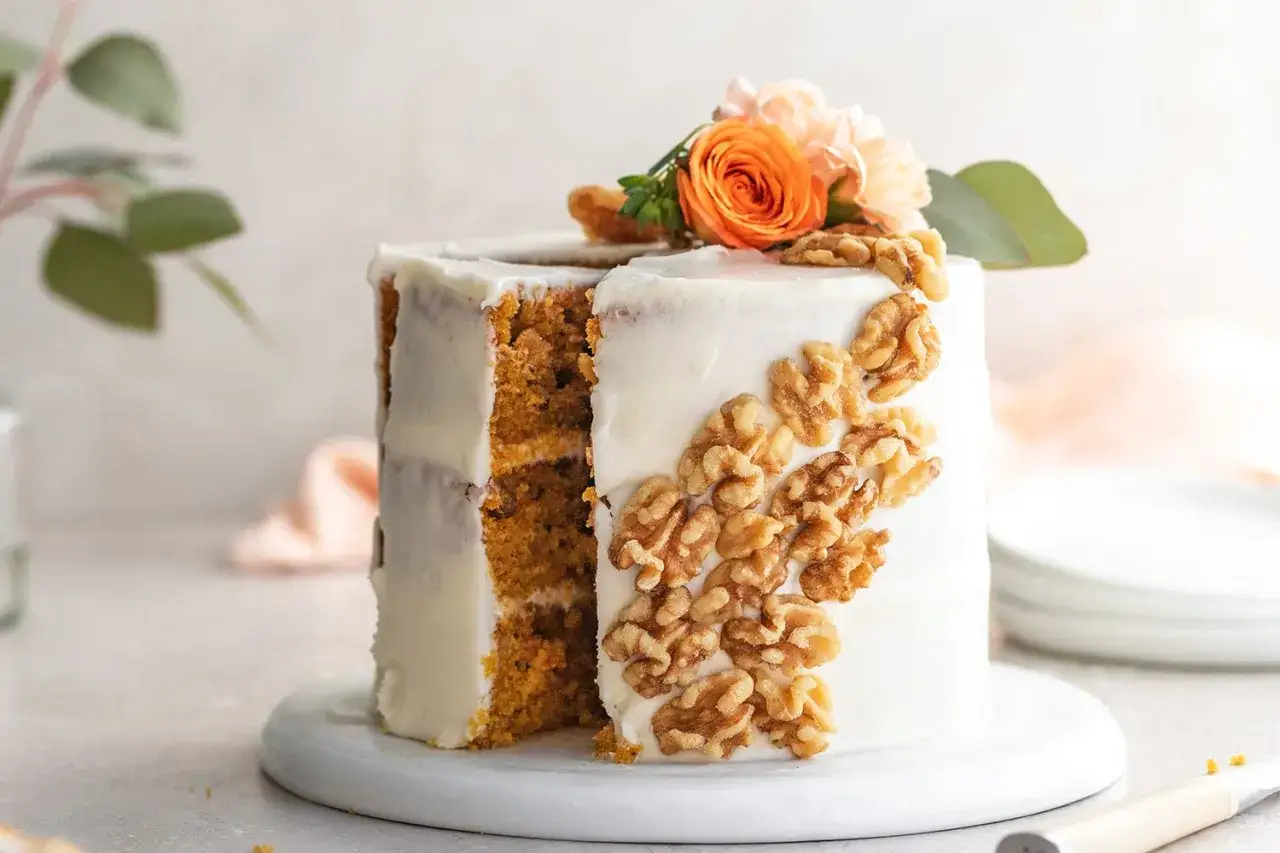 Tarta de zanahoria decorada con nueces y flores naranjas. Un corte revela sus capas esponjosas y el glaseado cremoso.