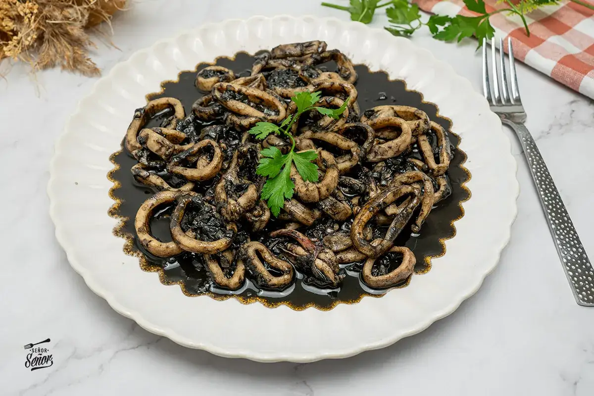 Calamares en su tinta, un plato tradicional español, servido en una fuente blanca con un tenedor y perejil fresco.