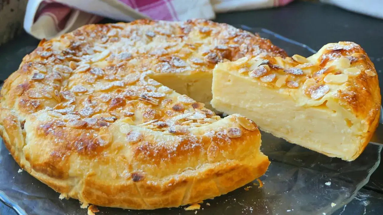 Tarta dorada con almendras laminadas y espolvoreada con azúcar glas. Una porción muestra su cremoso relleno, evocando el sabor del dulce de leche.