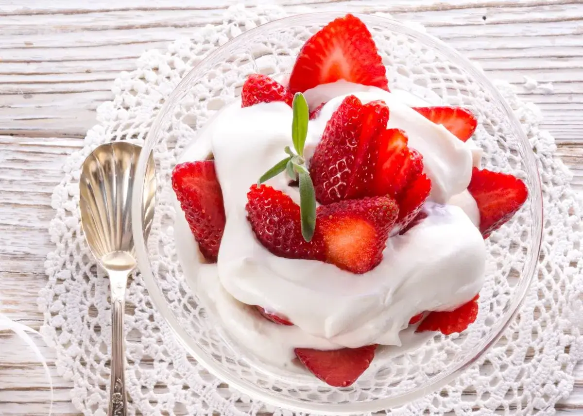 Fresas frescas cubiertas con abundante nata montada, servidas en un cuenco de cristal sobre un mantel de encaje.