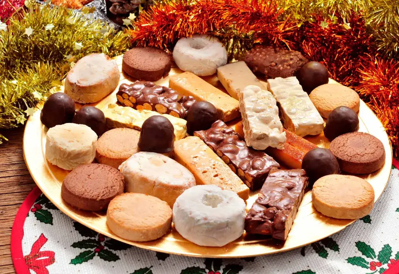 Variedad de postres navideños en una bandeja dorada: turrones, polvorones, mazapanes y bombones, adornados con espumillón festivo.