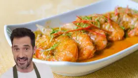 Un hombre sonriente presenta un plato de gambas al ajillo, con gambas jugosas en una salsa naranja, adornadas con finas tiras de chile rojo y hierbas verdes.