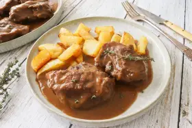 Dos tiernas porciones de pata de ternera guisada en su jugo, acompañadas de patatas rústicas fritas y una ramita de tomillo.
