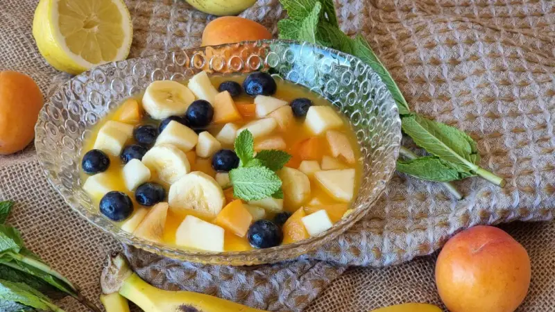 Deliciosa macedonia de frutas con plátano, arándanos, melocotón y manzana en un cuenco de cristal, decorada con menta.